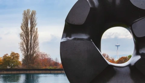 Black sculptural form with circular opening framing autumn foliage, waterway, and distant tower.