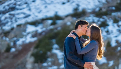 Couple kissing outdoors with snow-covered mountains and evergreen trees in soft golden hour light.