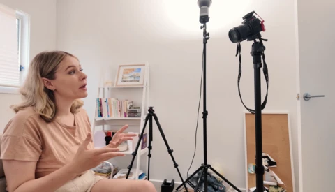 Woman sitting in a home studio setup, gesturing while speaking to camera during a recording session.