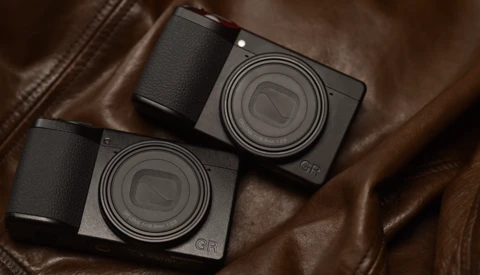 Two compact digital cameras with black bodies and textured grips positioned on brown leather fabric.