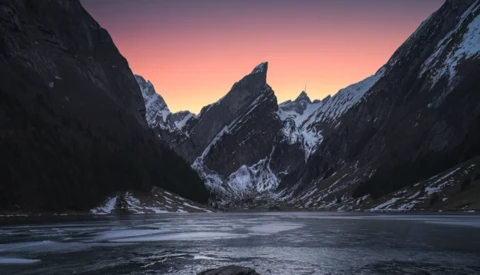Snow-covered mountains frame a frozen alpine lake at dawn, with pink and orange sky reflected in still water.