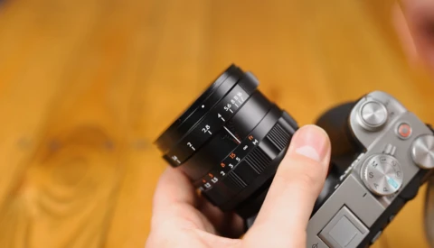 Hand holding a black telephoto lens next to a silver mirrorless camera body against a warm yellow background.