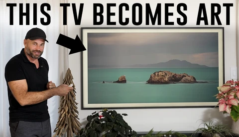Man in black polo shirt pointing to framed landscape photo displaying turquoise water and rocky islands.