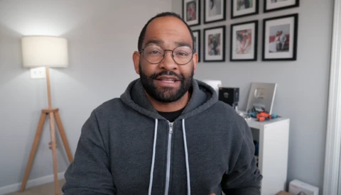 Man wearing glasses and dark hoodie smiling at camera in home office setting.
