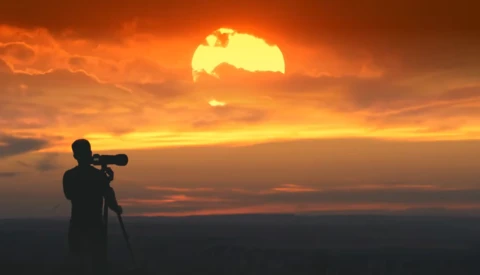 Silhouetted photographer with telephoto lens on tripod shooting a massive golden sunset.