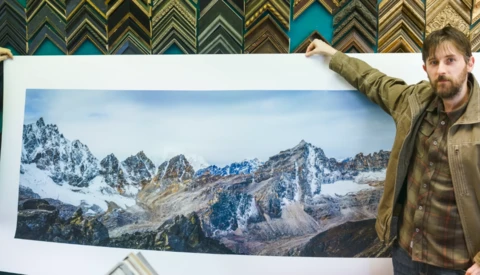 Photographer holding a large alpine landscape print featuring snow-capped peaks and rocky terrain.