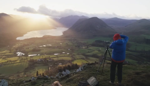 Photographer on mountainside with tripod photographing a lake valley at golden hour sunrise.