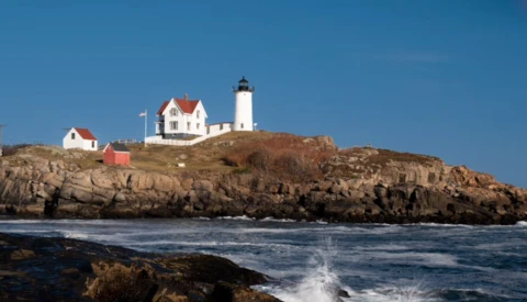 White lighthouse with red roof on rocky coastal headland under clear blue sky.