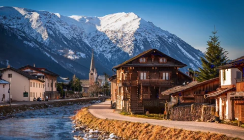 Snow-capped mountains rise behind a riverside Alpine village with wooden chalets and a church steeple.