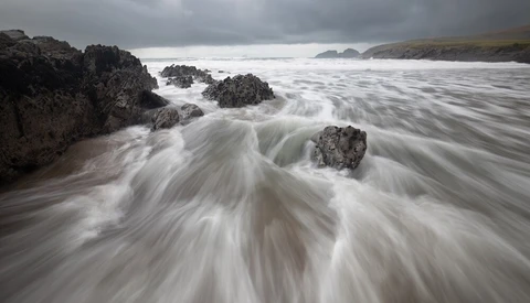 Silky seawater swirls around dark rocky outcrops on a sandy beach under overcast skies.