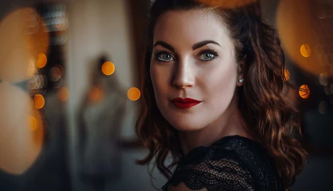 Woman with wavy dark hair and red lipstick looking over shoulder at camera with warm bokeh lights in background.