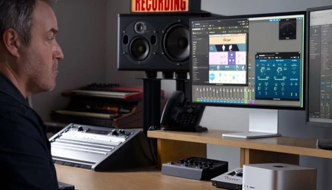 Music producer working at a recording studio desk with studio monitors, audio interface, and dual monitors displaying DAW software.