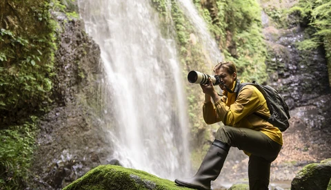 Photographer in yellow jacket crouching by moss-covered rocks, shooting a tall waterfall with a telephoto lens.