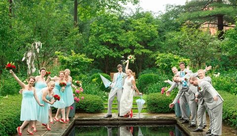 Wedding party posing on either side of a pond with the groom lifting the bride over the water.