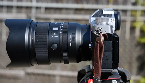 Mirrorless camera mounted on a tripod with a large telephoto lens attached, shown in profile against a blurred outdoor background.