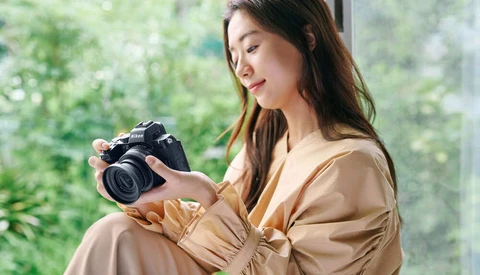 Woman holding a DSLR camera while seated by a bright window with green foliage visible outside.