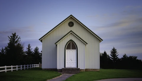 White wooden chapel with peaked roof and arched doorway at dusk, flanked by evergreen trees.