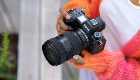 Photographer holding a Nikon DSLR camera with telephoto lens while wearing a bright pink and orange knit sweater.