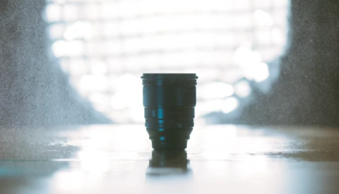 Dark telephoto lens standing upright on wet pavement with blurred bright background.