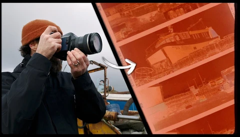 Photographer holding DSLR camera with telephoto lens, wearing brown beanie and dark jacket, positioned in front of orange industrial structure.