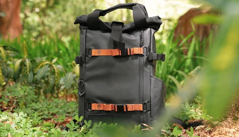Black roll-top backpack with orange straps positioned on a forest path surrounded by green vegetation.