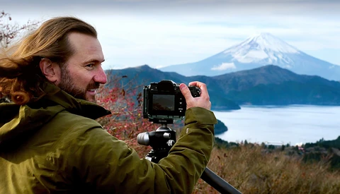 Photographer with long hair in olive jacket operating a mirrorless camera on tripod, framing a snow-capped volcanic peak across a lake.
