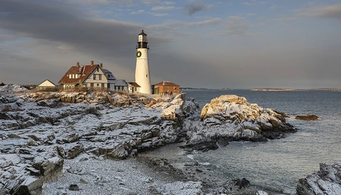 Portland Head Light lighthouse with keeper's house on rocky Maine coastline, snow-covered rocks in foreground, winter conditions.