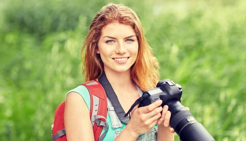 Young woman with reddish-blonde hair holding a professional DSLR camera with telephoto lens outdoors.