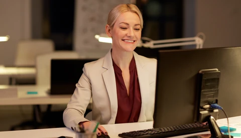 Professional woman at office desk with computer monitor and microphone, wearing blazer and burgundy top.