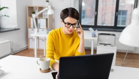 Person in yellow shirt wearing glasses, working at laptop at bright white desk with coffee cup.