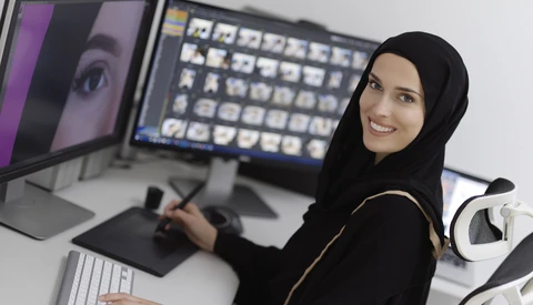 Woman at computer workstation with dual monitors and graphics tablet, wearing black hijab and smiling at camera.