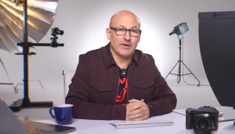 Man in maroon jacket speaking at desk in professional studio with lighting equipment.