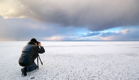 Photographer crouching on a salt flat with camera on tripod, facing dramatic cloudy sky at horizon.