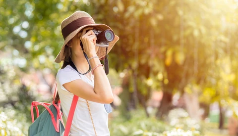 Young person wearing a hat and sunglasses looking through binoculars in a sunlit park setting.