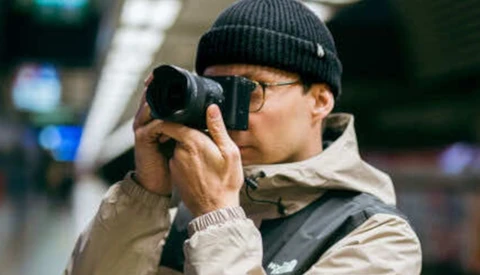 Photographer wearing glasses and a beanie holds a mirrorless camera up to his face while shooting in an indoor urban environment.