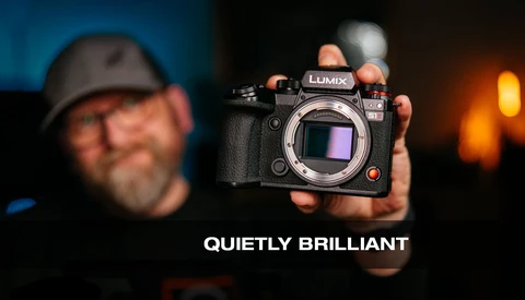 Photographer holding a Lumix mirrorless camera body toward the camera against a blurred background with warm lighting.