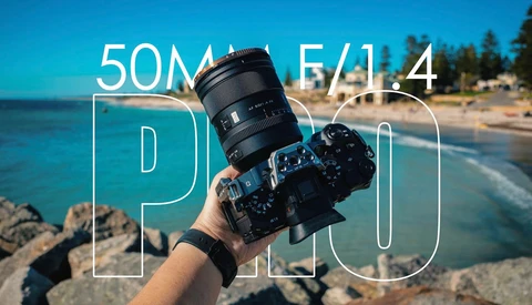 Photographer holding a camera with a 50mm f/1.4 lens at a coastal beach location with clear blue sky.