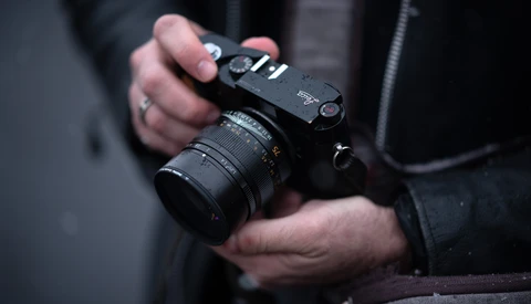 Photographer holding a mirrorless camera with attached lens and shoulder strap.