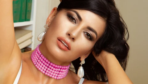 Woman with black wavy hair wearing pink beaded choker and pearl drop earrings, posed against neutral background.