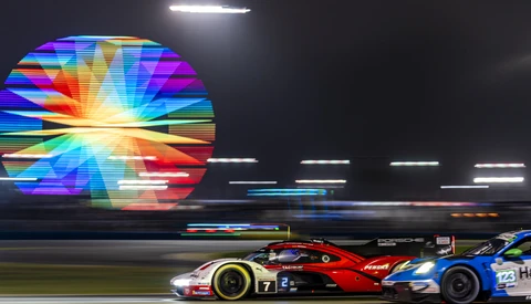 Red and blue race car on track at night with large colorful digital display in background.