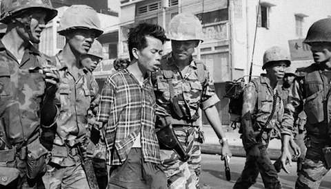 Armed soldiers escorting a civilian man in a plaid shirt through a street lined with buildings.