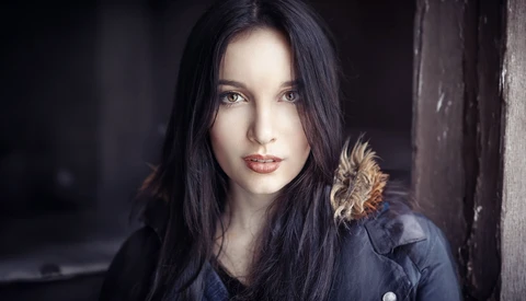 Woman with long black hair wearing dark clothing against a blurred dark background.