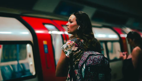 Woman with dark hair wearing a colorful floral jacket stands in a subway car with red doors and blue seating visible behind her.