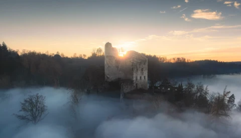 Medieval stone tower backlit by sunrise, surrounded by dense fog over a forested landscape.
