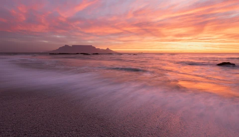 Sunrise over a coastal bay with a flat-topped mountain silhouetted against an orange and pink sky, waves washing onto the beach in the foreground.