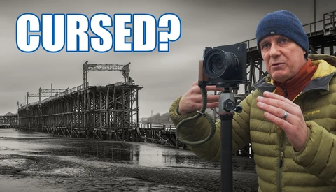 Photographer with camera and light meter standing before abandoned industrial steel structure.