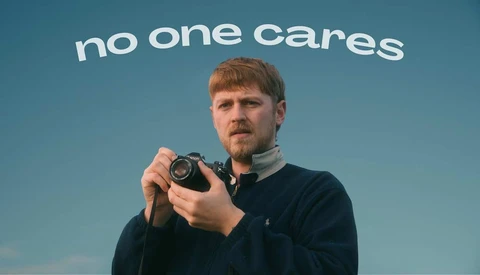 Man holding a vintage camera against a blue sky backdrop with "no one cares" text overhead.