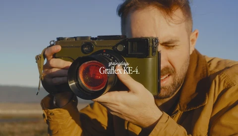 Photographer holding a Graflex KE-4 camera with olive-green body and red lens element against clear sky.