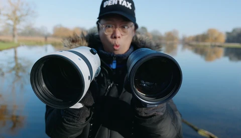 Photographer holding two telephoto lenses toward camera with wide-angle perspective distortion effect.