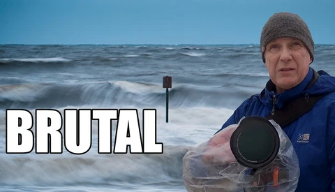 Photographer in winter gear holding waterproof camera equipment by the ocean.